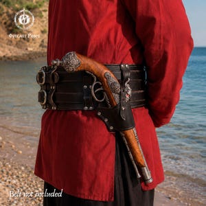 Op de afbeelding: Een bruin houten rekwisietpistool met gedetailleerde zilverkleurige metalen accenten op de handgreep en loop, bevestigd in een zwarte lederen holster. De holster zit vast aan een brede zwarte lederen riem met zilverkleurige gespen en klinknagels.