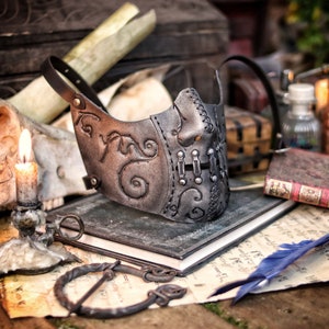 A silver and black leather mask with intricate designs and metal accents. The mask has a stitched seam down the center and is resting on a stack of books. Death Wizard Leather Mask M2 - Handcrafted Mask for Dark mages witches and powerful sorcerers with Intricate Details and Metallic Finish