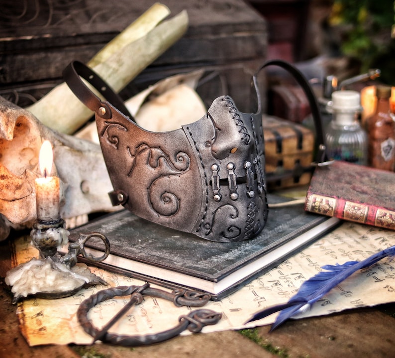 A silver and black leather mask with intricate designs and metal accents. The mask has a stitched seam down the center and is resting on a stack of books. Death Wizard Leather Mask M2 - Handcrafted Mask for Dark mages witches and powerful sorcerers with Intricate Details and Metallic Finish