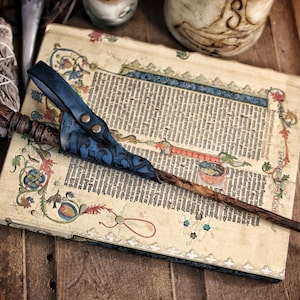 A blue embossed leather wand holster with aged texture rests over a decorated medieval manuscript while holding a long carved wand. The holster’s curved frog shape, riveted belt loop and hand dyed gradient give it a fantasy inspired look suited for wizard cosplay, witch costume, druid characters and larp wand holder accessories. The surrounding props enhance the mystical, scholarly aesthetic of the scene.