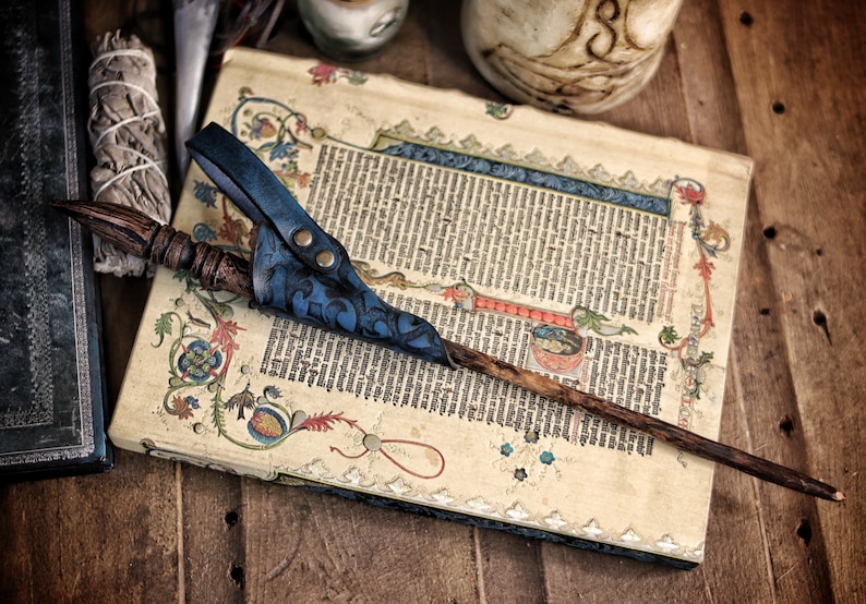 A blue embossed leather wand holster with aged texture rests over a decorated medieval manuscript while holding a long carved wand. The holster’s curved frog shape, riveted belt loop and hand dyed gradient give it a fantasy inspired look suited for wizard cosplay, witch costume, druid characters and larp wand holder accessories. The surrounding props enhance the mystical, scholarly aesthetic of the scene.