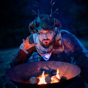 Fantasy druid costume with antler crown, oak leaves, and handcrafted Viking leather shoulder armor Sedrik, richly engraved with Celtic knots for LARP fantasy druid cosplay, magical shaman outfit or Nordic forest mage attire. The druid kneels by a campfire under a blue night sky, performing a ritual. Perfect for LARP, Celtic fantasy costume, druid cosplay, Viking fantasy warrior outfit, shaman costume, and medieval forest events or photoshoots. High-quality leather armor detail.