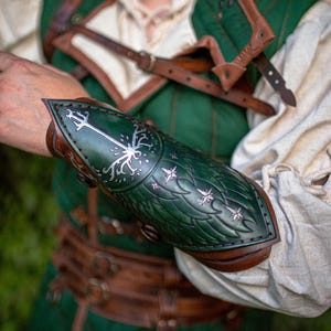 Op de afbeelding: Groene en bruine leren armbeschermer met een gestileerd boom- en sterrenontwerp. De armbeschermer maakt deel uit van een kostuum, met een bijpassend groen vest en bruine leren riemen. De tekst "Outcast Props" is zichtbaar.