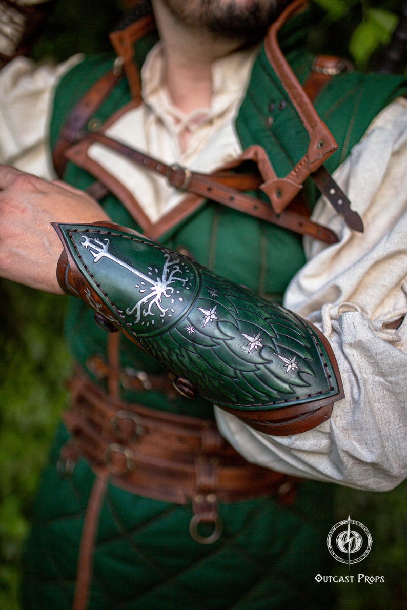 Op de afbeelding: Groene en bruine leren armbeschermer met een gestileerd boom- en sterrenontwerp. De armbeschermer maakt deel uit van een kostuum, met een bijpassend groen vest en bruine leren riemen. De tekst "Outcast Props" is zichtbaar.