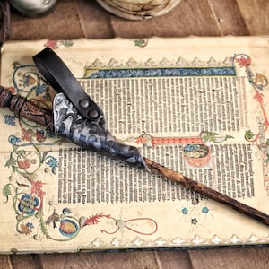 A silver black embossed leather wand holster hangs from its belt loop while holding a long wooden wand, placed over an antique illuminated manuscript with ornate borders and medieval lettering. The holster’s curved frog shape and textured finish evoke handcrafted wizard gear. A sage bundle and rustic props surround it, creating an immersive fantasy setting suited for larp wand holder needs, wizard cosplay accessories and renaissance faire magic outfits.