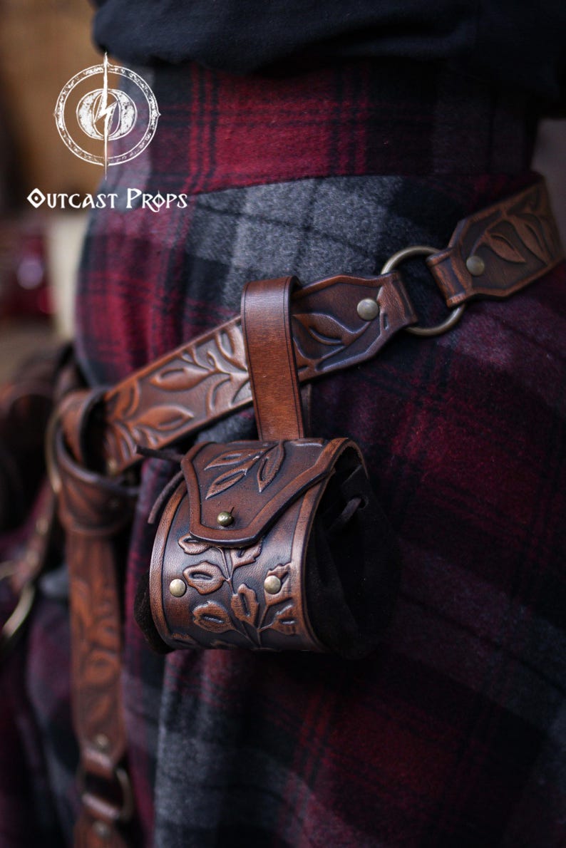 A brown floral embossed leather coin pouch hangs from a matching carved belt worn over a red and grey plaid skirt. The botanical design and aged leather finish give it a natural elven or druidic style. Perfect for renaissance faire or renfaire outfits, the pouch offers a soft suede body for carrying coins, dice or tiny essentials. A practical accessory for witches, herbalists, nobles or fantasy LARP characters seeking detailed leather gear.