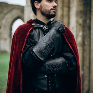 A fantasy LARP warrior wearing dark leather clothing and a red velvet cloak shows off black Yggdrasil leather bracers embossed with Norse world tree details. These long bracers, perfect for viking costume, leather armor, or medieval cosplay, offer both protection and character depth. Ideal for renaissance fairs, Geralt cosplay, or as female leather armor accessories. Model stands in front of a ruined stone archway, crossing arms with solemn expression. Yggdrasil