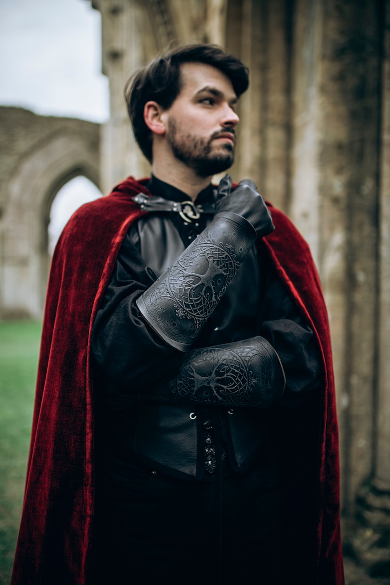 A fantasy LARP warrior wearing dark leather clothing and a red velvet cloak shows off black Yggdrasil leather bracers embossed with Norse world tree details. These long bracers, perfect for viking costume, leather armor, or medieval cosplay, offer both protection and character depth. Ideal for renaissance fairs, Geralt cosplay, or as female leather armor accessories. Model stands in front of a ruined stone archway, crossing arms with solemn expression. Yggdrasil
