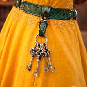 May include: A green leather belt with a leaf design and silver metal hardware. The belt holds a set of antique-style keys. The background is a yellow dress. The text "Belt not included" is visible.