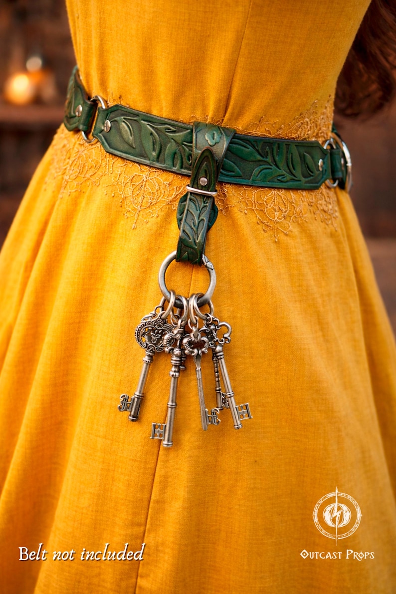 May include: A green leather belt with a leaf design and silver metal hardware. The belt holds a set of antique-style keys. The background is a yellow dress. The text "Belt not included" is visible.