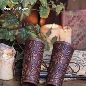 Two aged brown floral leather bracers stand together on a rustic tabletop surrounded by ivy leaves and candlelight. The renaissance faire bracer design features embossed petals and leaves that create a refined floral arm guard ideal for fantasy outfits. This scene highlights the handcrafted texture, making each piece a fitting elf cosplay accessory or fae costume accessory for larp events and medieval inspired looks. Fae costume, ren faire, druid larp, leaf design, floral design.