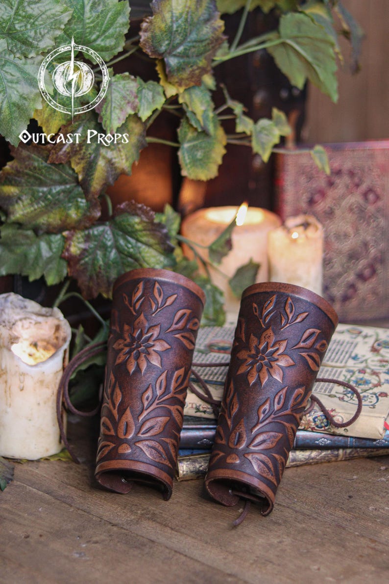 Two aged brown floral leather bracers stand together on a rustic tabletop surrounded by ivy leaves and candlelight. The renaissance faire bracer design features embossed petals and leaves that create a refined floral arm guard ideal for fantasy outfits. This scene highlights the handcrafted texture, making each piece a fitting elf cosplay accessory or fae costume accessory for larp events and medieval inspired looks. Fae costume, ren faire, druid larp, leaf design, floral design.