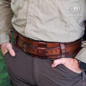 Close side view of the aged brown bushcraft leather belt worn over outdoor clothing, showing the adjustable inner strap, wide outer frame, brass buckle, stitched edges and layered construction. Handmade leather utility belt designed for bushcraft gear, camping, hiking, survival setups, tactical belt use, military inspired costumes, steampunk outfits, LARP characters and medieval fantasy clothing. The belt offers a rugged look for everyday wear and outdoor events.