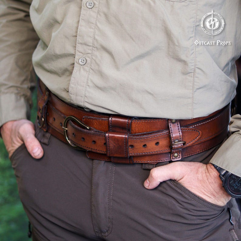 Close side view of the aged brown bushcraft leather belt worn over outdoor clothing, showing the adjustable inner strap, wide outer frame, brass buckle, stitched edges and layered construction. Handmade leather utility belt designed for bushcraft gear, camping, hiking, survival setups, tactical belt use, military inspired costumes, steampunk outfits, LARP characters and medieval fantasy clothing. The belt offers a rugged look for everyday wear and outdoor events.