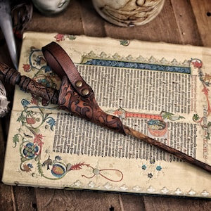 A rich brown embossed leather wand holster with ornate patterns holds a carved wooden wand across an illuminated manuscript filled with medieval text and colorful decorations. Its belt loop and brass toned rivets show artisan craftsmanship. The warm tones and rustic background evoke wizard cosplay, druid or witch costume styles. Ideal for renaissance faire outfits, larp wand holder accessories and magic wand belt holster displays.