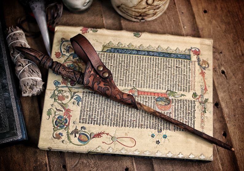 A rich brown embossed leather wand holster with ornate patterns holds a carved wooden wand across an illuminated manuscript filled with medieval text and colorful decorations. Its belt loop and brass toned rivets show artisan craftsmanship. The warm tones and rustic background evoke wizard cosplay, druid or witch costume styles. Ideal for renaissance faire outfits, larp wand holder accessories and magic wand belt holster displays.