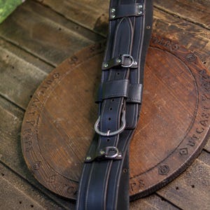May include: A dark brown leather belt featuring multiple adjustable straps, silver-toned metal buckles, and reinforced rivets. The belt has a wide, structured design with visible stitching along the edges and a central vertical strap configuration.