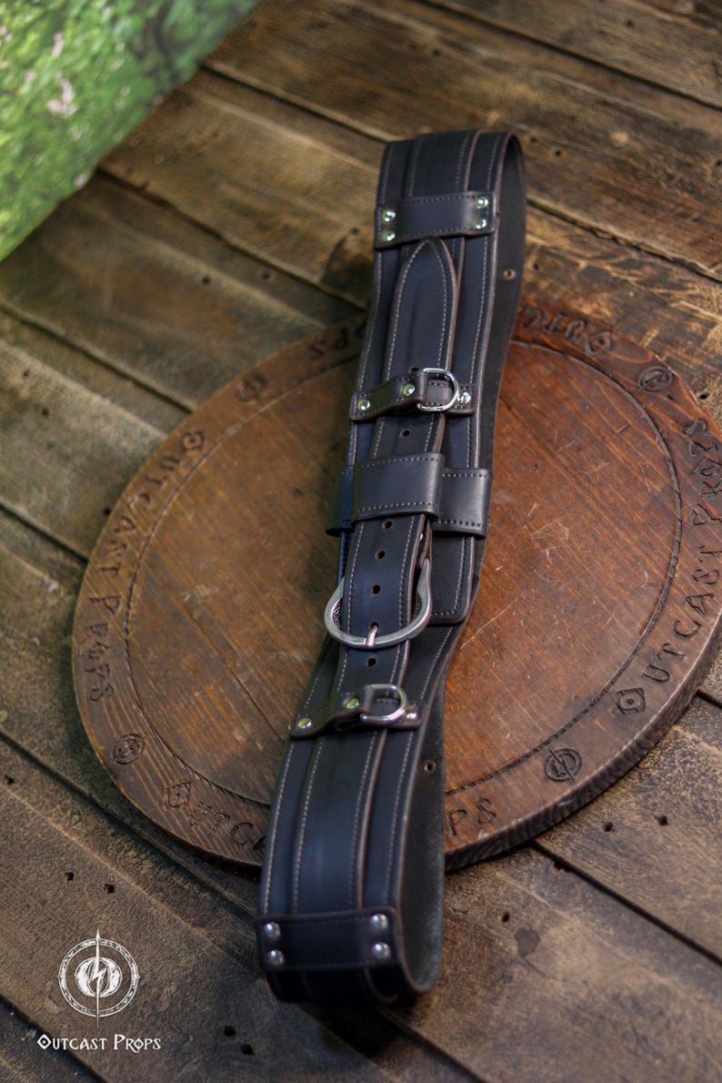 May include: A dark brown leather belt featuring multiple adjustable straps, silver-toned metal buckles, and reinforced rivets. The belt has a wide, structured design with visible stitching along the edges and a central vertical strap configuration.