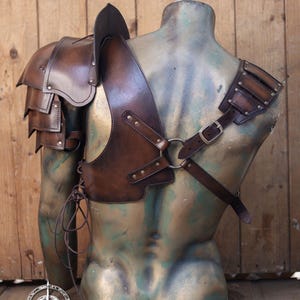Pu&ograve; includere: Set di spallacci e imbracatura in pelle marrone. L'armatura presenta una protezione per le spalle a strati, una cinghia pettorale con anello metallico e chiusura con fibbia. Il testo "SHOULDER PAULDRON + HARNESS" &egrave; nella parte superiore dell'immagine.