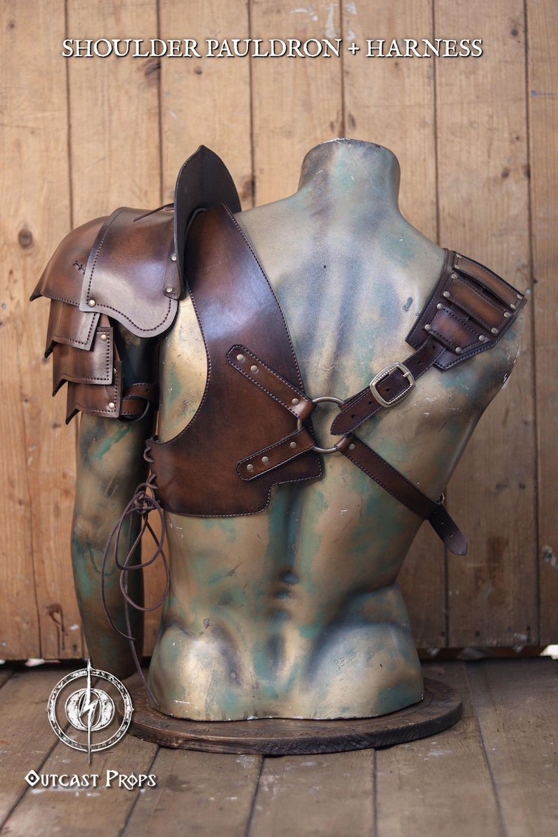 Pu&ograve; includere: Set di spallacci e imbracatura in pelle marrone. L'armatura presenta una protezione per le spalle a strati, una cinghia pettorale con anello metallico e chiusura con fibbia. Il testo "SHOULDER PAULDRON + HARNESS" &egrave; nella parte superiore dell'immagine.