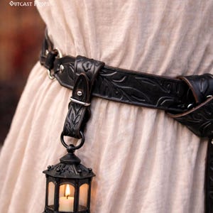 May include: Black leather belt with a decorative leaf pattern and a hanging lantern. The lantern is black with clear glass panels and a lit candle inside. The text "Outcast Props" is visible.