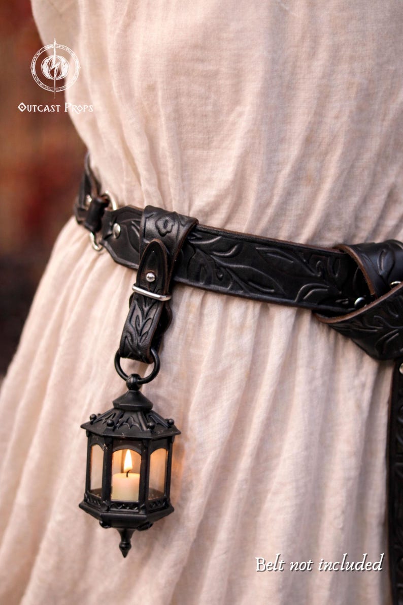 May include: Black leather belt with a decorative leaf pattern and a hanging lantern. The lantern is black with clear glass panels and a lit candle inside. The text "Outcast Props" is visible.