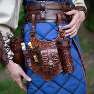 Puede incluir: Un cinturón utilitario de cuero marrón con múltiples bolsillos y un soporte para viales. El cinturón se lleva sobre una prenda acolchada azul. El cuero está intrincadamente trabajado con patrones decorativos.