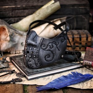 A black leather mask with intricate silver metal accents and a floral design. The mask is on a dark surface with a blue feather and a book. Death Wizard Leather Mask M2 - Handcrafted Mask for Dark mages witches and powerful sorcerers with Intricate Details and Metallic Finish