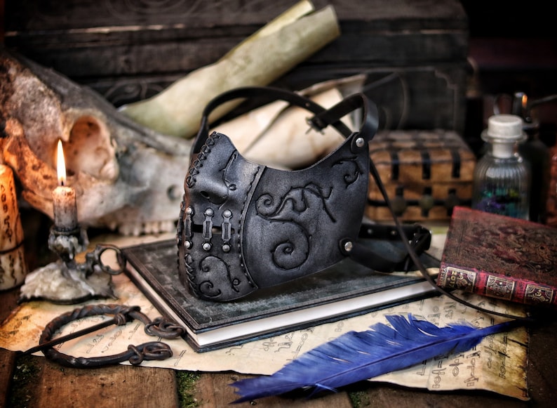 A black leather mask with intricate silver metal accents and a floral design. The mask is on a dark surface with a blue feather and a book. Death Wizard Leather Mask M2 - Handcrafted Mask for Dark mages witches and powerful sorcerers with Intricate Details and Metallic Finish