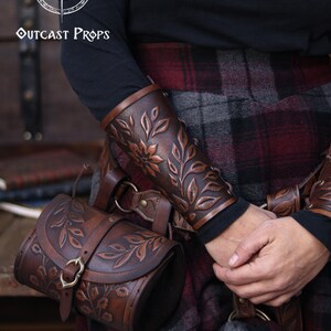 Two aged brown floral leather bracers are worn on crossed arms above a richly textured renaissance faire belt with a matching embossed pouch. The floral arm guard design features carved leaves and petals with deep highlights that emphasize the artisan finish. This larp bracer set fits naturally into medieval styled clothing, offering a fantasy cosplay accessory suitable for druids, elves or ranger characters. Warm browns and soft lighting enhance the handcrafted detail. Fae druid