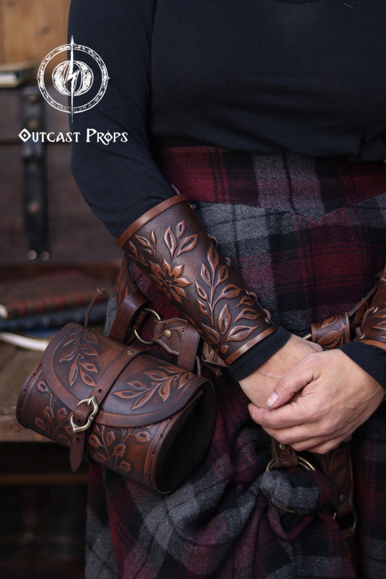 Two aged brown floral leather bracers are worn on crossed arms above a richly textured renaissance faire belt with a matching embossed pouch. The floral arm guard design features carved leaves and petals with deep highlights that emphasize the artisan finish. This larp bracer set fits naturally into medieval styled clothing, offering a fantasy cosplay accessory suitable for druids, elves or ranger characters. Warm browns and soft lighting enhance the handcrafted detail. Fae druid