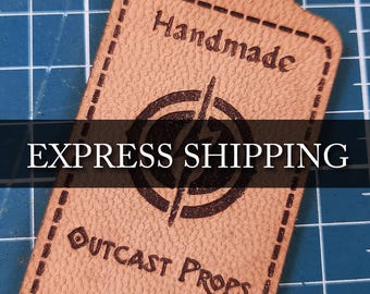 Könnte beinhalten: Ein braunes Leder-Etikett mit einem genähten Rand. Das Etikett hat ein Logo mit einem Blitz in einem Kreis und den Text "Outcast Props" und "outcastprops.com". Das Etikett trägt außerdem die Aufschrift "Handmade" und "EXPRESS SHIPPING".