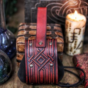 Handcrafted leather coin pouch in aged red vegetable-tanned leather, photographed front-on on a wooden table. The pouch features deep geometric embossing inspired by Celtic and dwarf motifs, a suede body that expands for coins, and a sturdy belt loop at the back. Brass rivets and hand-dyed aging give it a rugged yet refined look. Styled with candles, glass bottles and a carved wooden chest, this leather coin pouch suits LARP, Viking, Celtic and fantasy costumes, Renaissance fairs,
