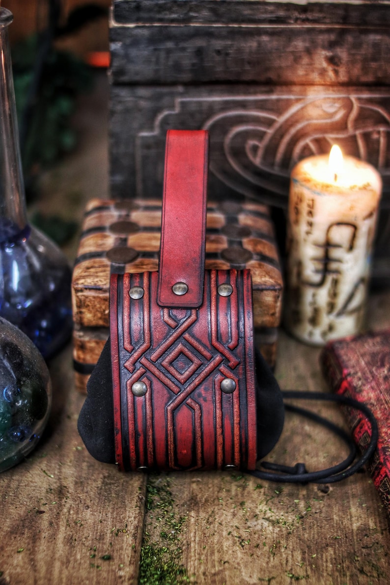 Handcrafted leather coin pouch in aged red vegetable-tanned leather, photographed front-on on a wooden table. The pouch features deep geometric embossing inspired by Celtic and dwarf motifs, a suede body that expands for coins, and a sturdy belt loop at the back. Brass rivets and hand-dyed aging give it a rugged yet refined look. Styled with candles, glass bottles and a carved wooden chest, this leather coin pouch suits LARP, Viking, Celtic and fantasy costumes, Renaissance fairs,