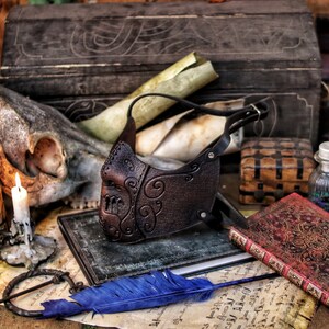 May include: A brown leather mask with intricate scrollwork designs. The mask is on a table with a skull, candles, and old books.