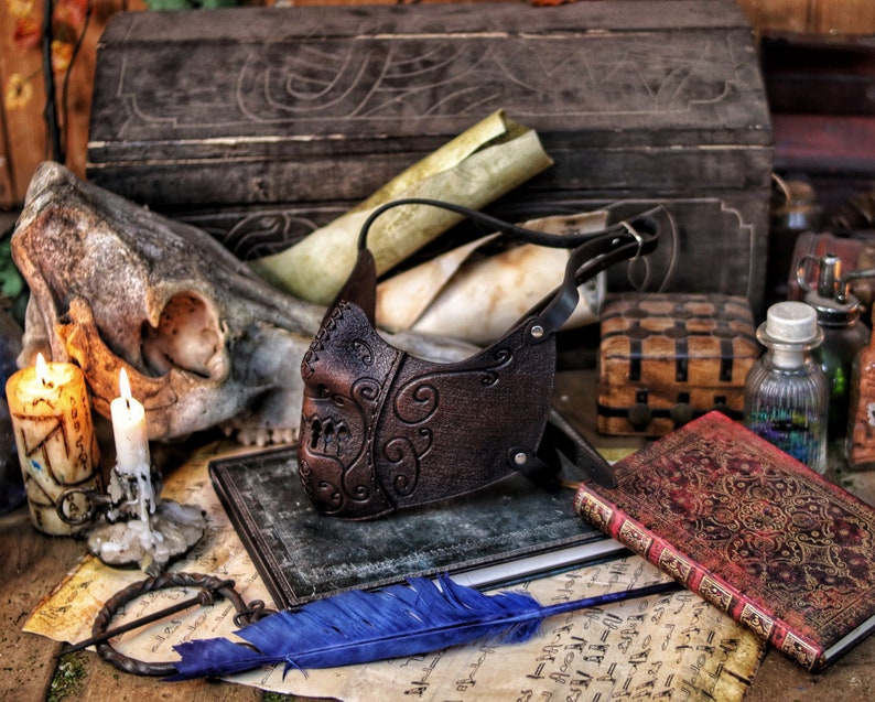 May include: A brown leather mask with intricate scrollwork designs. The mask is on a table with a skull, candles, and old books.