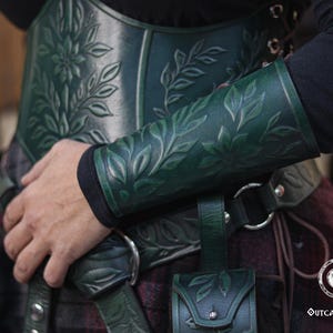 Close up of an aged green floral leather bracer with detailed embossed leaves, worn with a matching green corset for a renaissance faire costume. The renaissance faire bracer curves naturally around the forearm, making it ideal as an elf cosplay accessory or floral arm guard. Texture, shading and artisan dye work are clearly visible, showing a handcrafted larp bracer perfect for fae and woodland characters. Fae costume, ren faire, druid larp, leaf design, floral design.