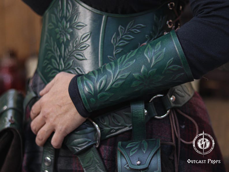 Close up of an aged green floral leather bracer with detailed embossed leaves, worn with a matching green corset for a renaissance faire costume. The renaissance faire bracer curves naturally around the forearm, making it ideal as an elf cosplay accessory or floral arm guard. Texture, shading and artisan dye work are clearly visible, showing a handcrafted larp bracer perfect for fae and woodland characters. Fae costume, ren faire, druid larp, leaf design, floral design.