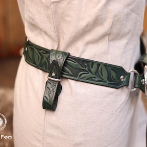 May include: Dark green leather belt with a leaf pattern. The belt has a buckle and a strap with a silver metal ring. The belt is worn over a light-colored garment. The belt is a decorative accessory.