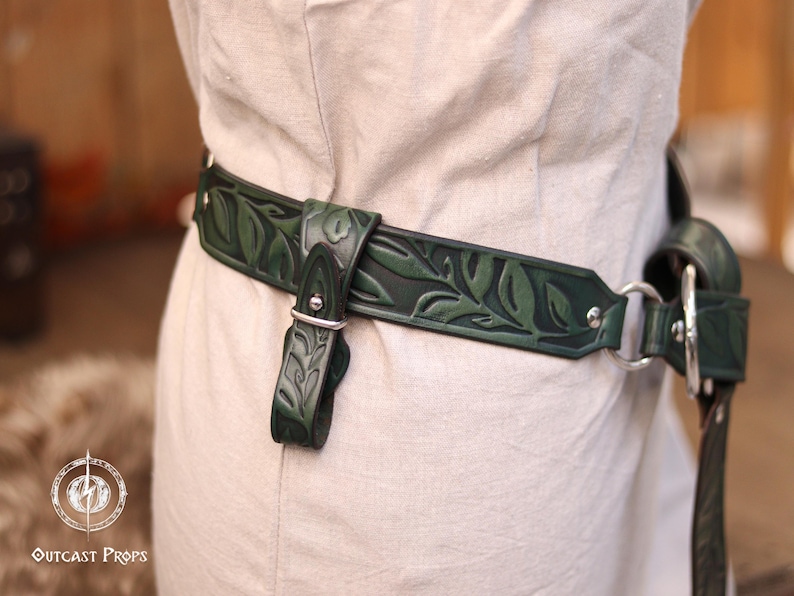 May include: Dark green leather belt with a leaf pattern. The belt has a buckle and a strap with a silver metal ring. The belt is worn over a light-colored garment. The belt is a decorative accessory.