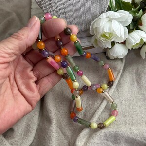 May include: A colorful beaded necklace featuring a variety of gemstone beads in shades of pink, orange, green, yellow, and purple. The necklace has gold-colored spacers and is displayed on a neutral background.