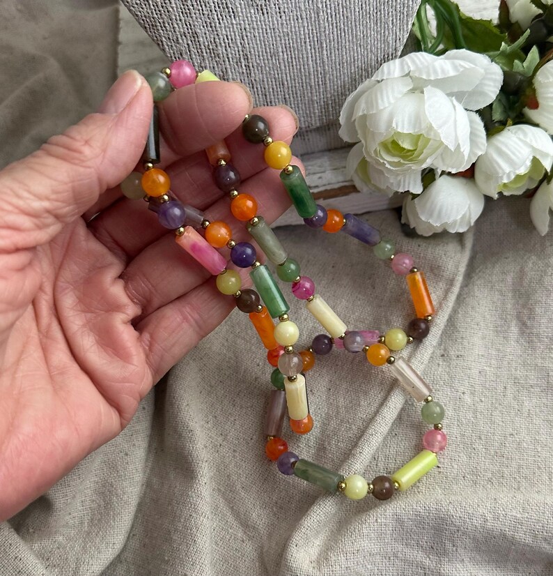 May include: A colorful beaded necklace featuring a variety of gemstone beads in shades of pink, orange, green, yellow, and purple. The necklace has gold-colored spacers and is displayed on a neutral background.