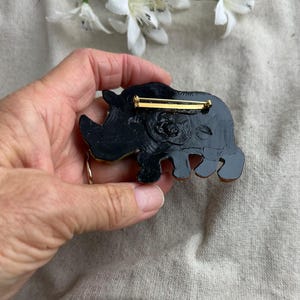 May include: A black and gold rhino brooch with a gold pin closure. The rhino is detailed with a textured surface and a gold horn. The brooch is a unique accessory for jackets, bags, or hats.