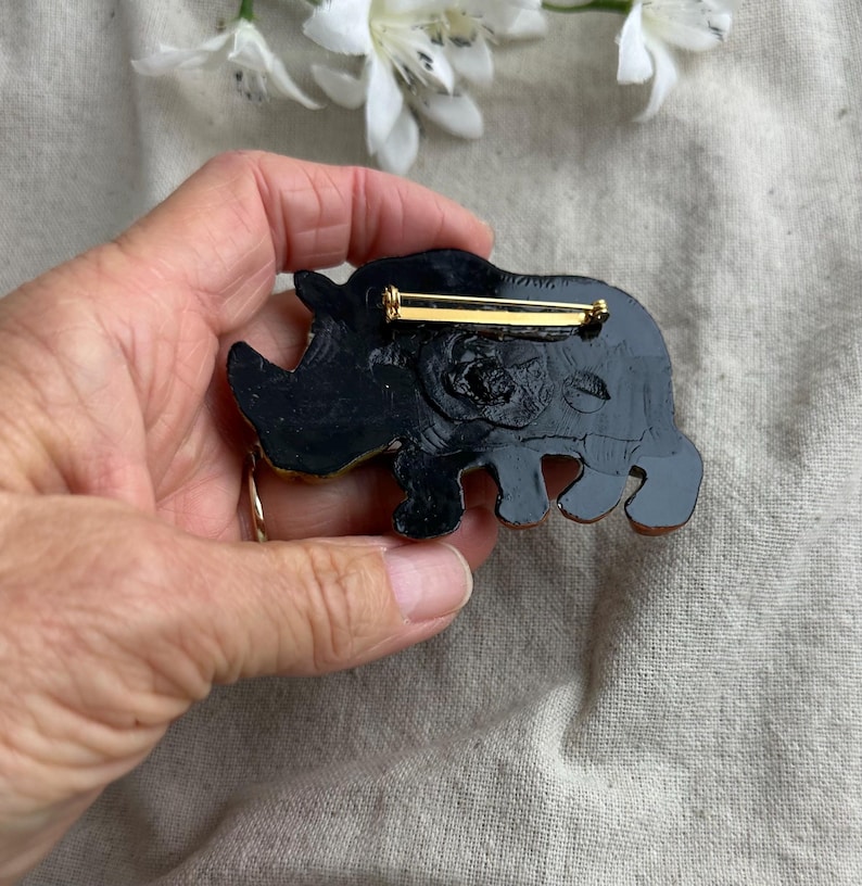 May include: A black and gold rhino brooch with a gold pin closure. The rhino is detailed with a textured surface and a gold horn. The brooch is a unique accessory for jackets, bags, or hats.