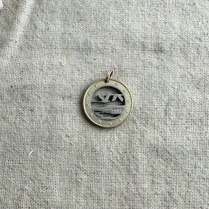 May include: A gold-colored metal pendant with a cutout design of two birds flying over a wave. The pendant has a star-shaped border and the year "2002" inscribed on it.