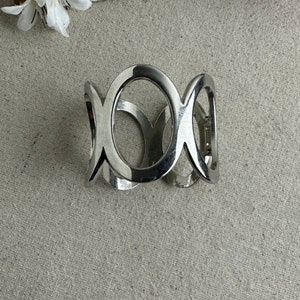 May include: A silver-toned cuff bracelet with an open design featuring three connected oval shapes. The bracelet has a hinged closure and a polished finish, reflecting light. The jewelry is displayed on a neutral-toned surface.