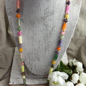 May include: A colorful necklace with a variety of gemstone beads in shades of orange, pink, green, yellow, and purple. The necklace is displayed on a mannequin bust with a decorative crown in the background. The necklace is a long style.