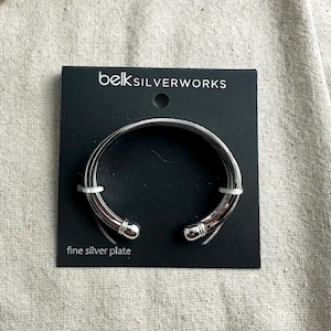 May include: A silver-plated cuff bracelet on a black Belk Silverworks card. The bracelet has a rounded, open design with a smooth, shiny finish. The card reads "fine silver plate".
