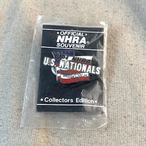 May include: A black and white pin with a red, white, and blue American flag design. The pin reads "1987 NHRA U.S. NATIONALS INDIANAPOLIS INDIANA" and "OFFICIAL NHRA SOUVENIR COLLECTORS EDITION".
