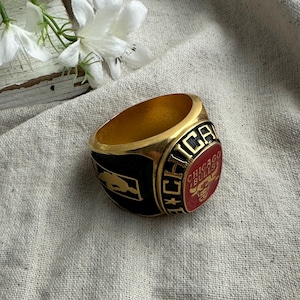 May include: A gold-colored ring with a black and red design. The ring features the words "CHICAGO BULLS" and a logo. The ring is set against a neutral background with white flowers.
